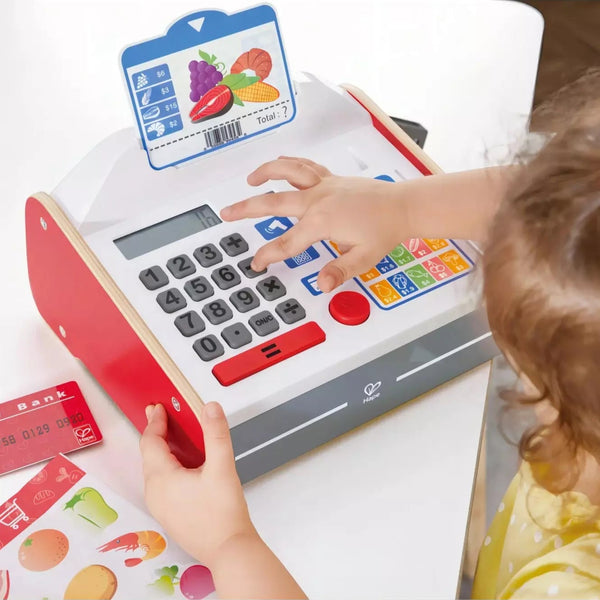 Child playing with a red and white toy cash register, featuring a scanner with light and sound effects, coins, and paper money.