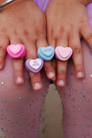 A set of colorful chunky heart rings, each with a sweet message on a finger. The rings are displayed against a backdrop of a sparkling, heart-patterned leotard.