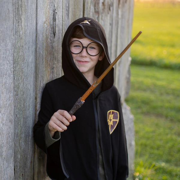 Child in a black robe with a Harry Potter-themed emblem, holding a wand, against a wooden wall.