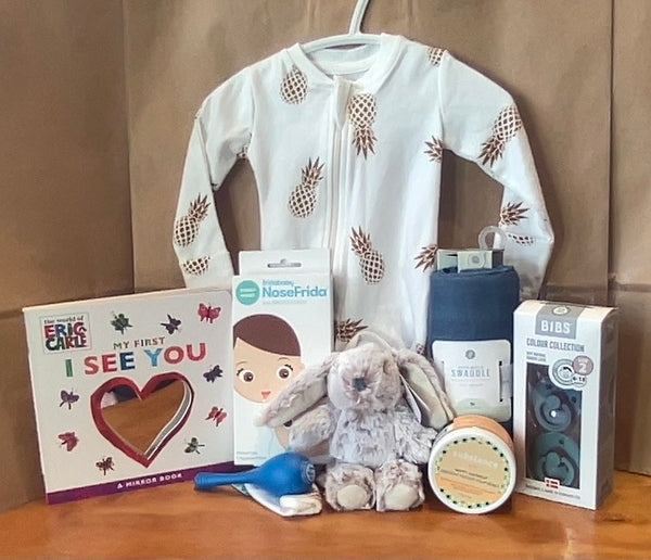 A collection of baby items displayed on a table, including a white onesie with a pattern, a blue and white maraca, a brown and white board book, a gray and blue soft toy, a white and blue pacifier, a white and blue rattle toy, and a green and white container.